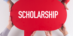 A red speech bubble sign with the word "Scholarship" held by multiple hands, symbolizing financial aid opportunities for students.
