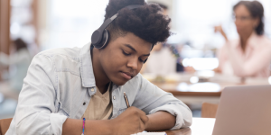 A young student wearing headphones and studying in a library, taking notes from a laptop.