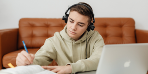 A student wearing headphones, studying at home with a laptop and taking notes.