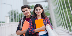 Two university students smiling while holding notebooks, standing on a modern university campus.