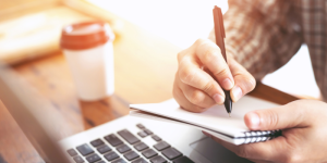 A student taking handwritten notes while studying Turkish online with a laptop and coffee