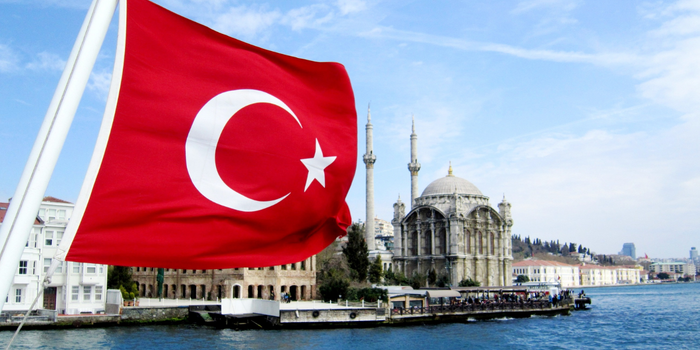Turkish flag waving near a mosque by the sea, symbolizing national identity and culture.