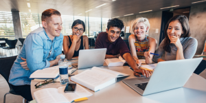 A diverse group of university students studying together around a table with laptops and books.