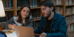 Two students studying together in a quiet library with bookshelves in the background.