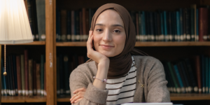 A smiling female student wearing a hijab, sitting in a university library surrounded by books.